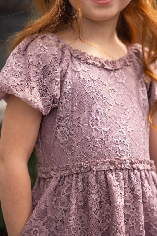 A young girl wears the Ivy City Co Mini Kiara Dress in Hyssop Lace, featuring dusty purple lace with short sleeves and floral patterns. She stands outdoors, her long red hair flowing and her face partially visible.