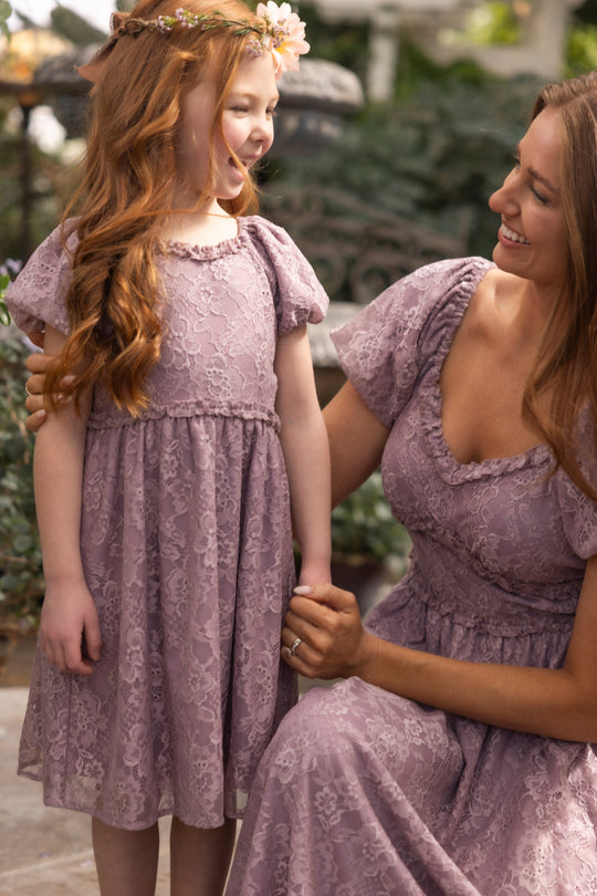 A woman and a young girl, both in Ivy City Co’s Mini Kiara Dress in Hyssop Lace, smile outdoors. The girl, with long red hair and a flower crown, holds hands with the kneeling woman in these charming matching party dresses.