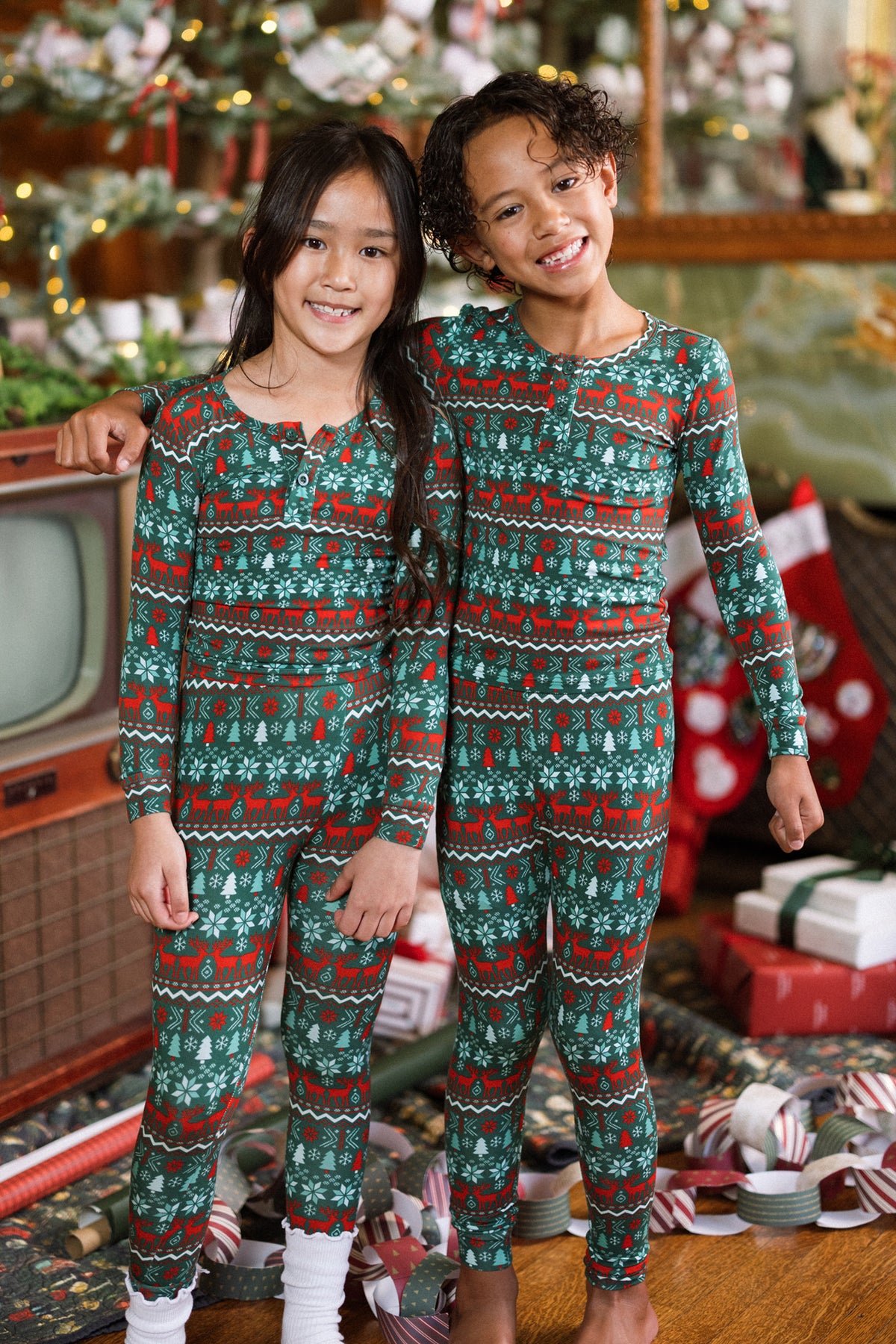Two children in matching Kids So Soft Pajama Set in Alpine Noel by Ivy City Co smile together in a festive room with a Christmas tree, wrapped gifts, stockings, and a vintage TV in the background.