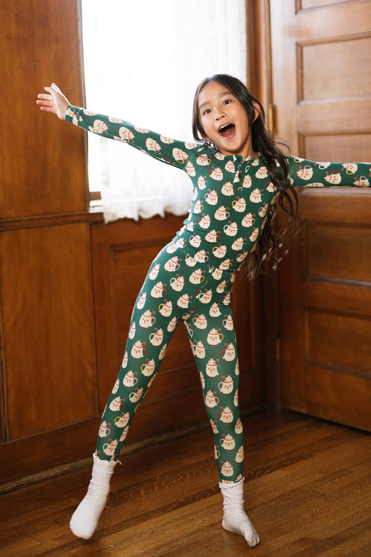 A young girl beams with joy, arms wide, in the Kids So Soft Pajama Set in Very Merry Santa by Ivy City Co—her cozy look complete with white socks and long dark hair, standing on a wooden floor in a warmly lit room.