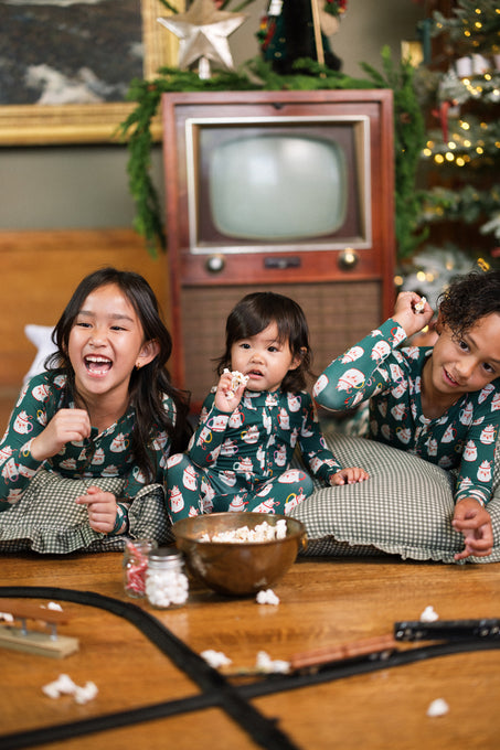 Three kids in the Kids So Soft Pajama Set in Very Merry Santa by Ivy City Co sit on the floor, smiling and eating popcorn near a Christmas tree, with a toy train and scattered popcorn adding festive cheer.