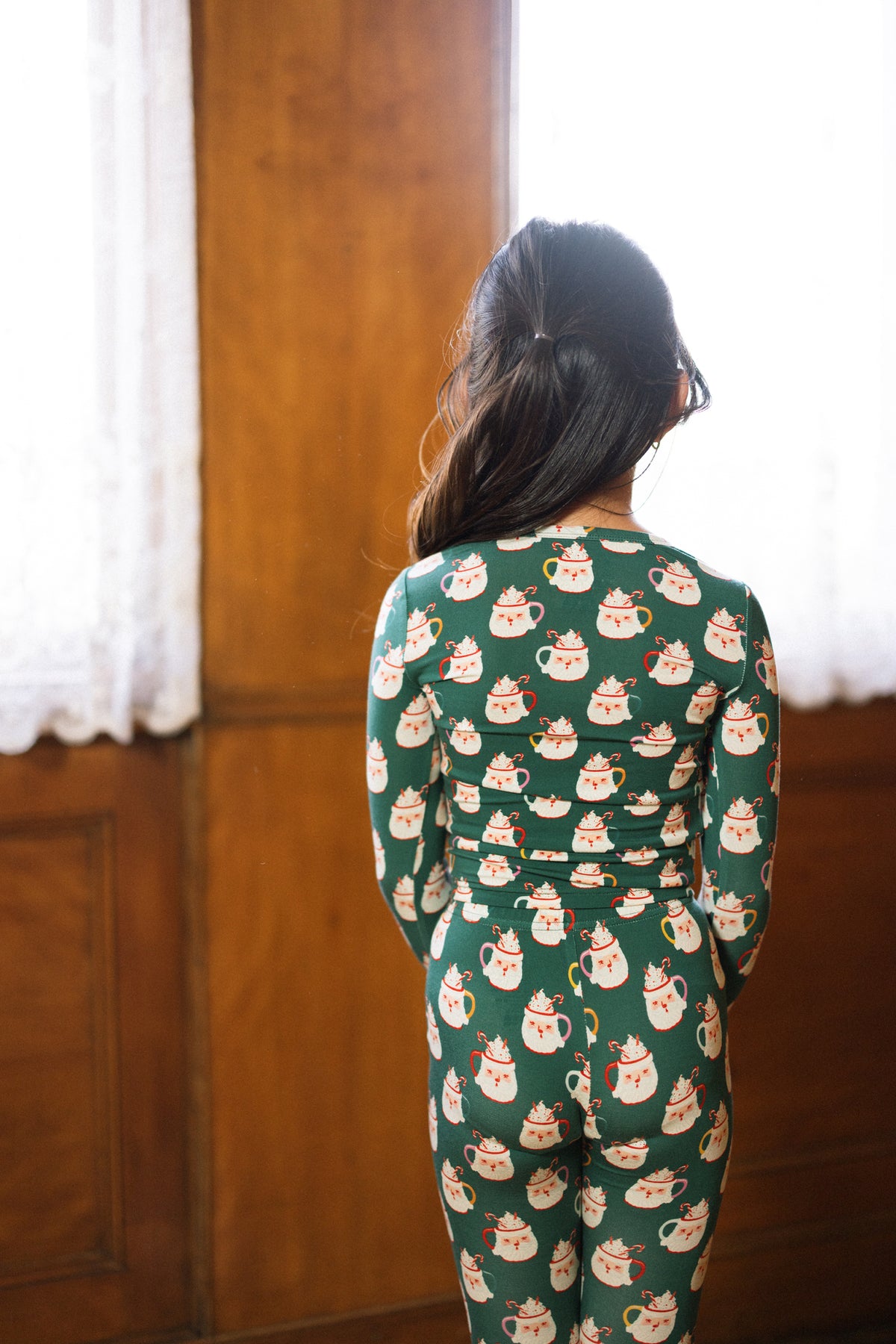 A girl with long dark hair wears the Kids So Soft Pajama Set in Very Merry Santa by Ivy City Co, standing by a sunlit window with lace curtains and wooden panels.