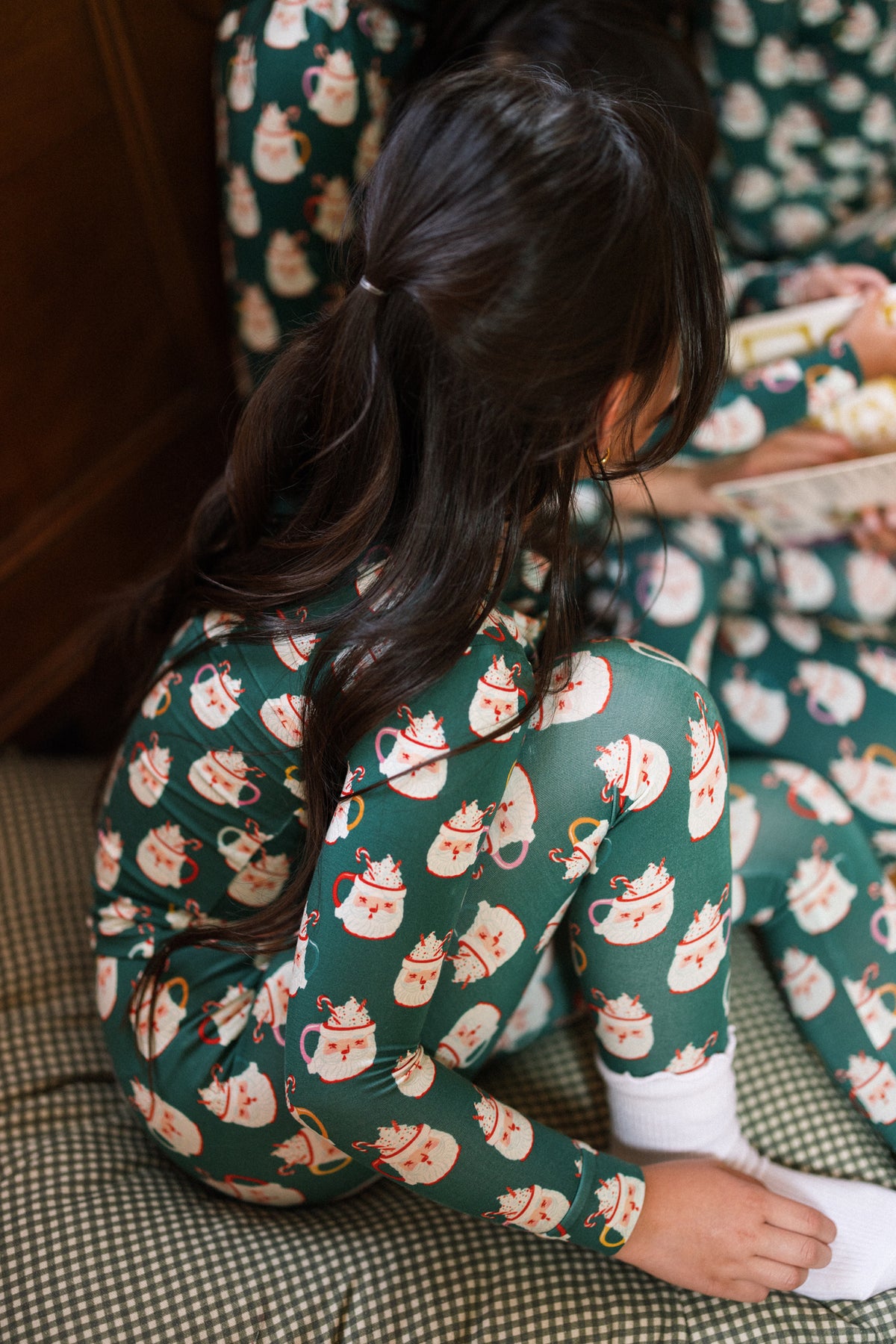 A child with long dark hair sits cross-legged on a plaid cushion, wearing the Ivy City Co Kids So Soft Pajama Set in Very Merry Santa, facing away in festive family matching pajamas.