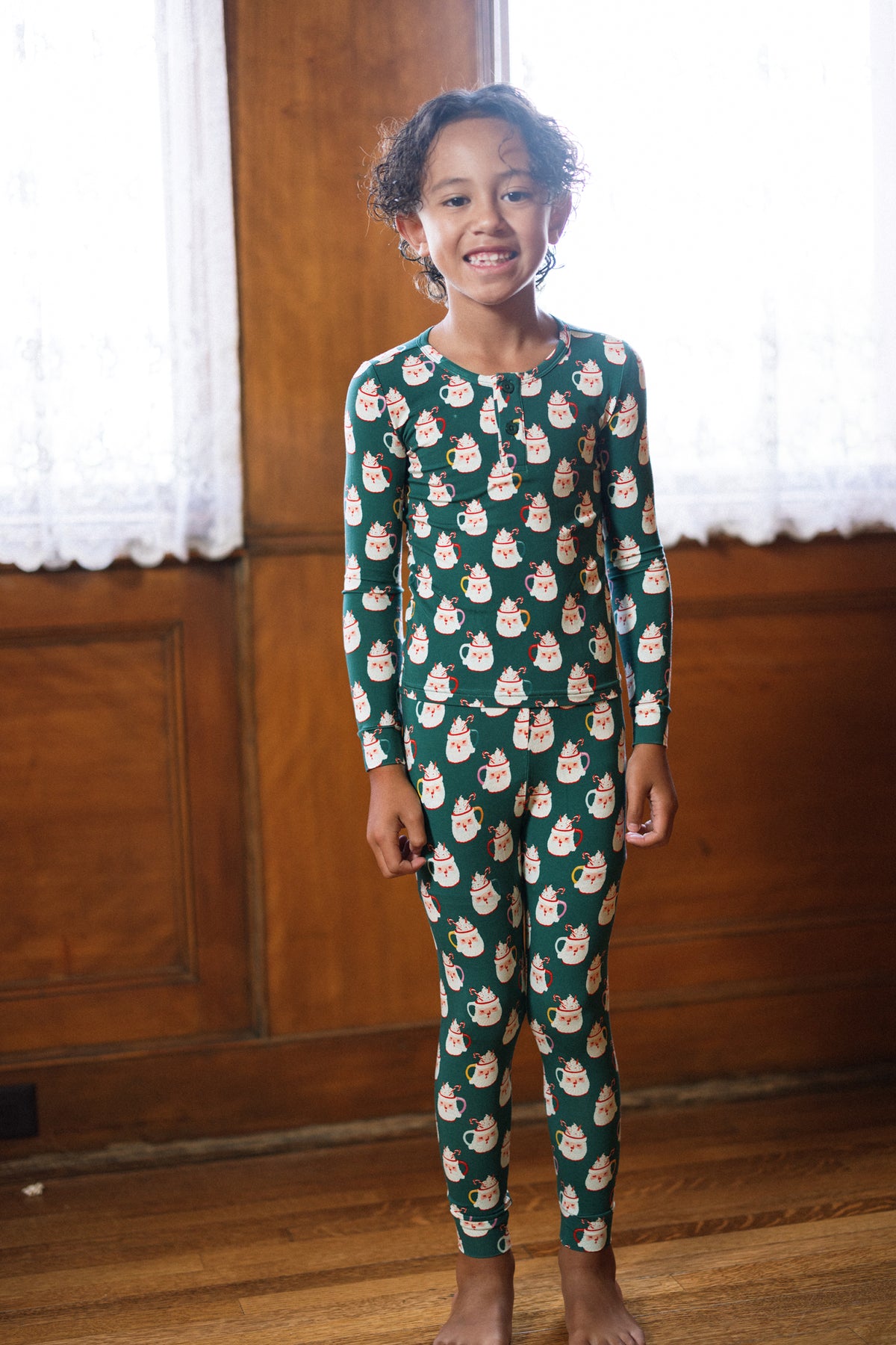 A smiling child stands barefoot indoors wearing the Ivy City Co Kids So Soft Pajama Set in Very Merry Santa, featuring matching green pajamas with Santa Claus print, in front of wood paneling and lace curtains.