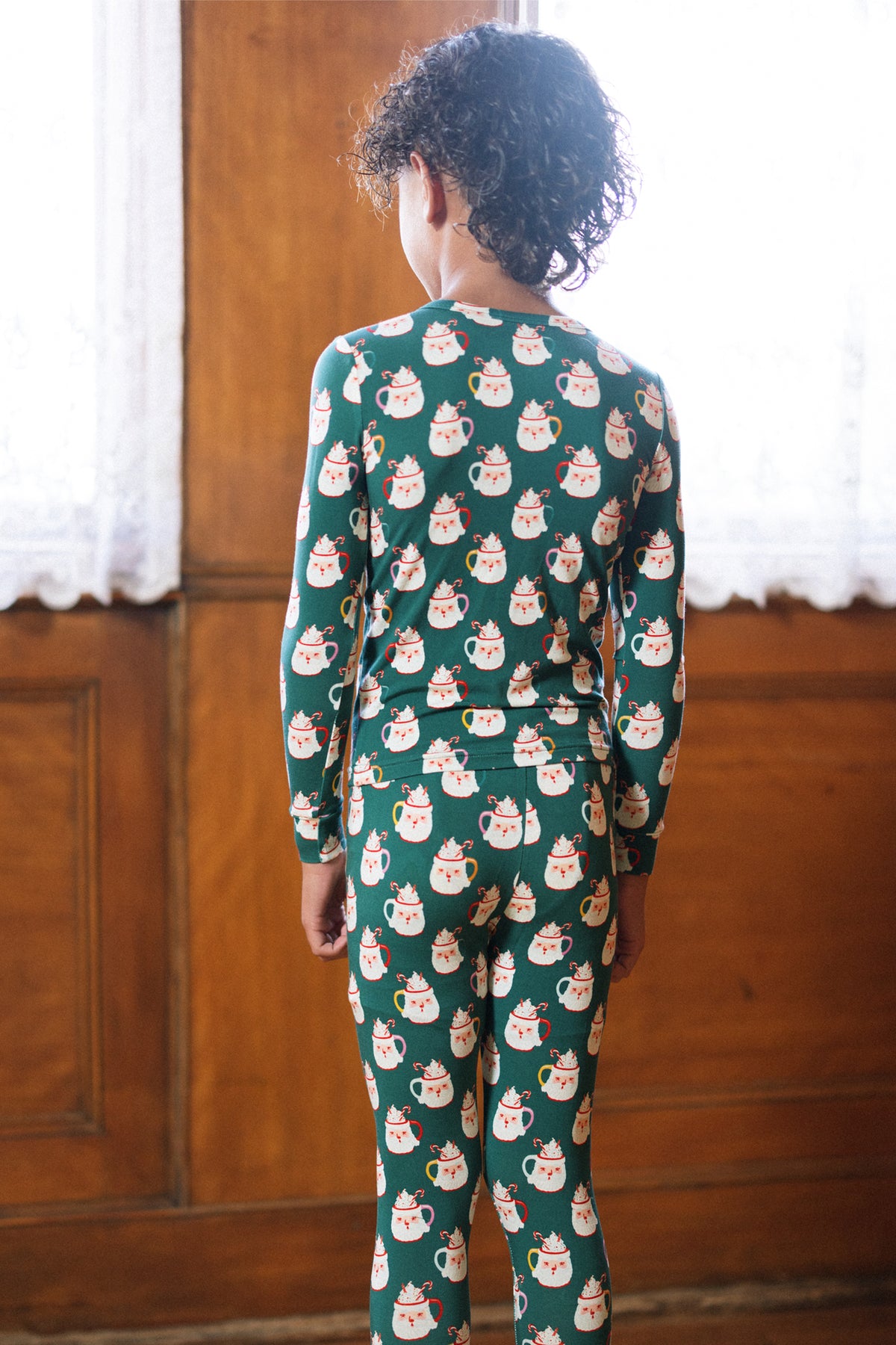 A child with curly hair stands facing away, wearing the Kids So Soft Pajama Set in Very Merry Santa by Ivy City Co. The scene features wooden paneling and white lace curtains.