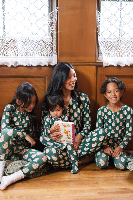 A woman and three children in Ivy City Co’s Kids So Soft Pajama Set in Very Merry Santa sit on a wood floor by a window. The smallest child holds a book as they all smile and laugh together.