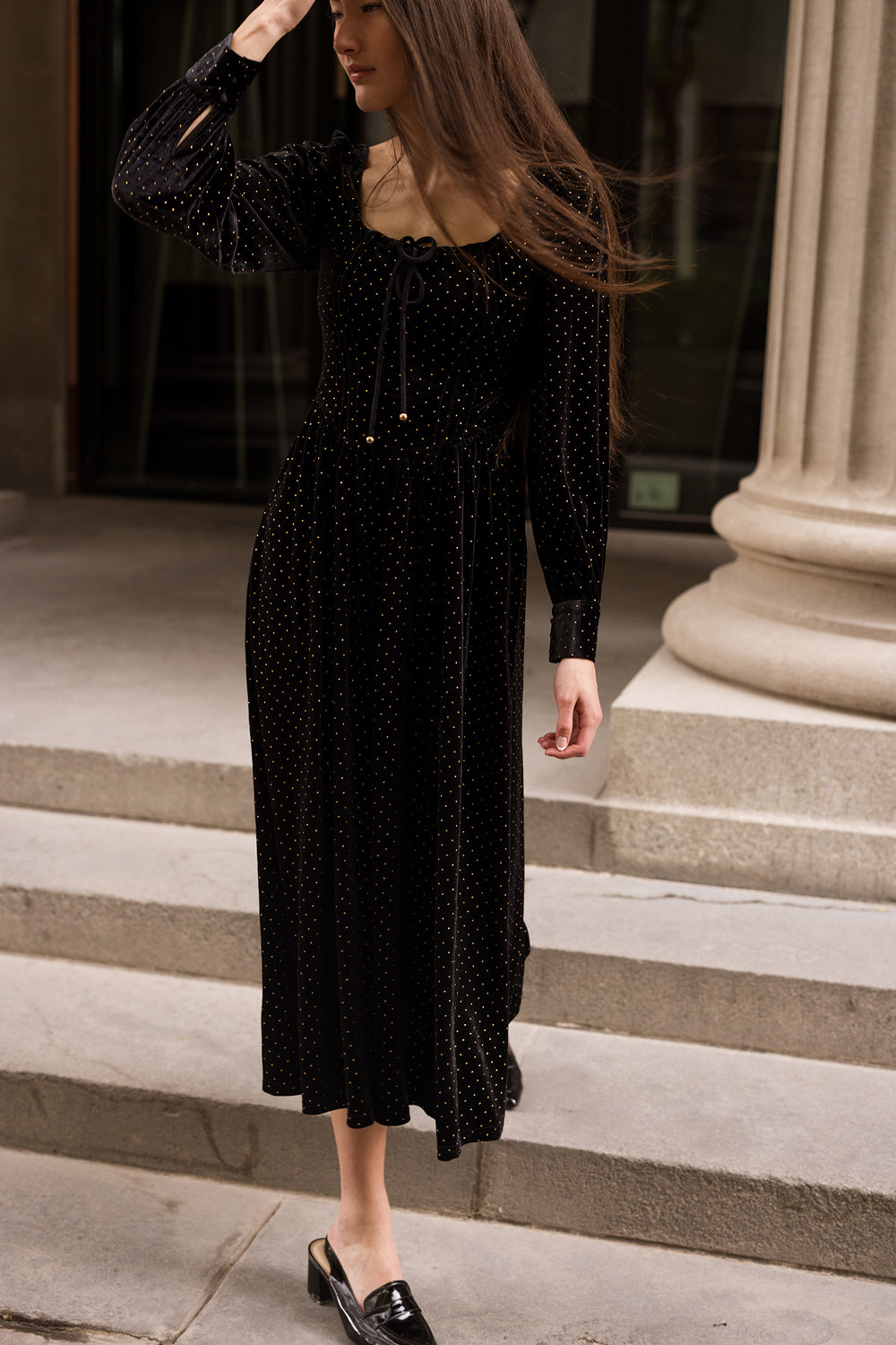 A woman wearing the Ivy City Co Kingston Dress in Black, styled with black loafers, stands on stone steps near a large column. Her long hair is down and she touches her head with one hand.