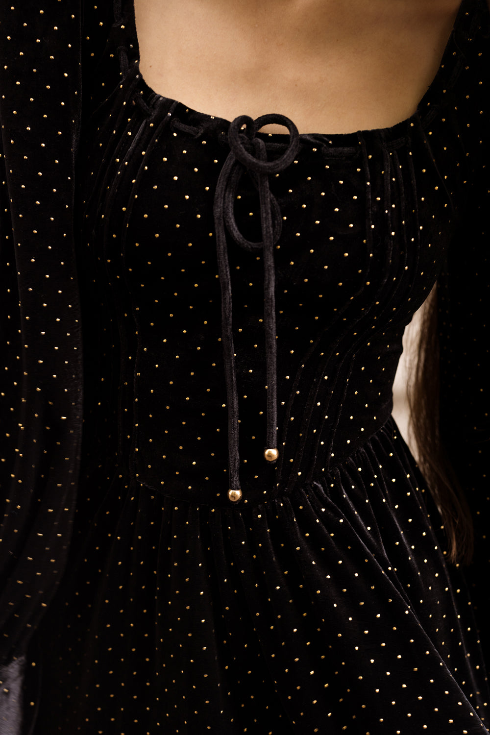 Close-up of the Ivy City Co Kingston Dress in Black, a velvet dress adorned with small gold polka dots and featuring a tie detail at the neckline, worn by a person with bare shoulders visible.
