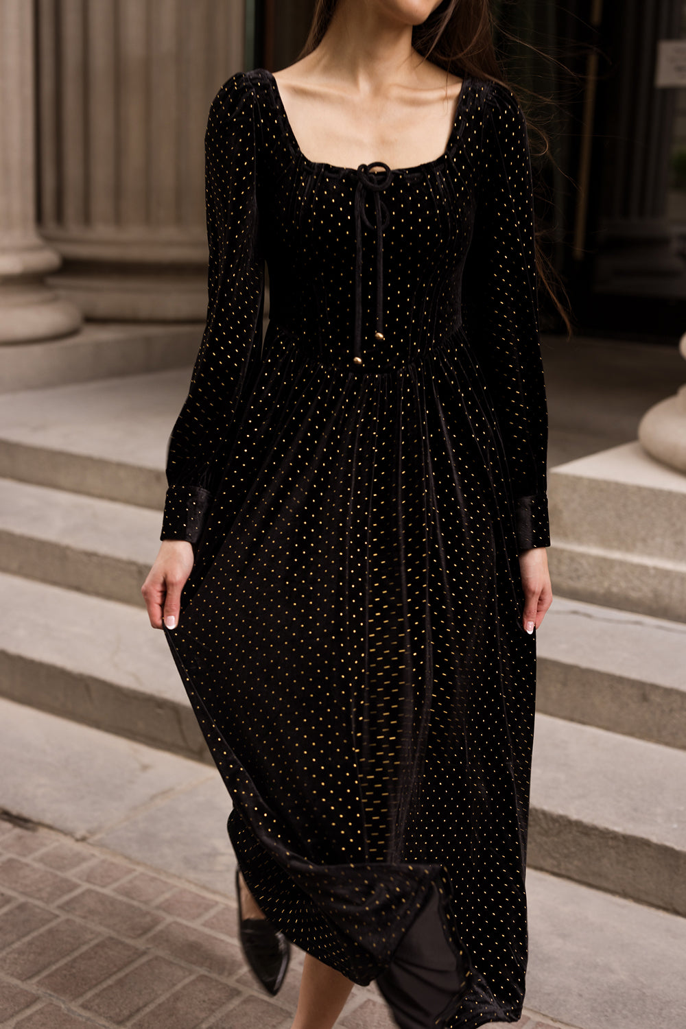 A woman with long brown hair wears the Ivy City Co Kingston Dress in Black, a long-sleeved velvet dress featuring gold polka dots, as she stands on stone steps framed by columns and an arched doorway.