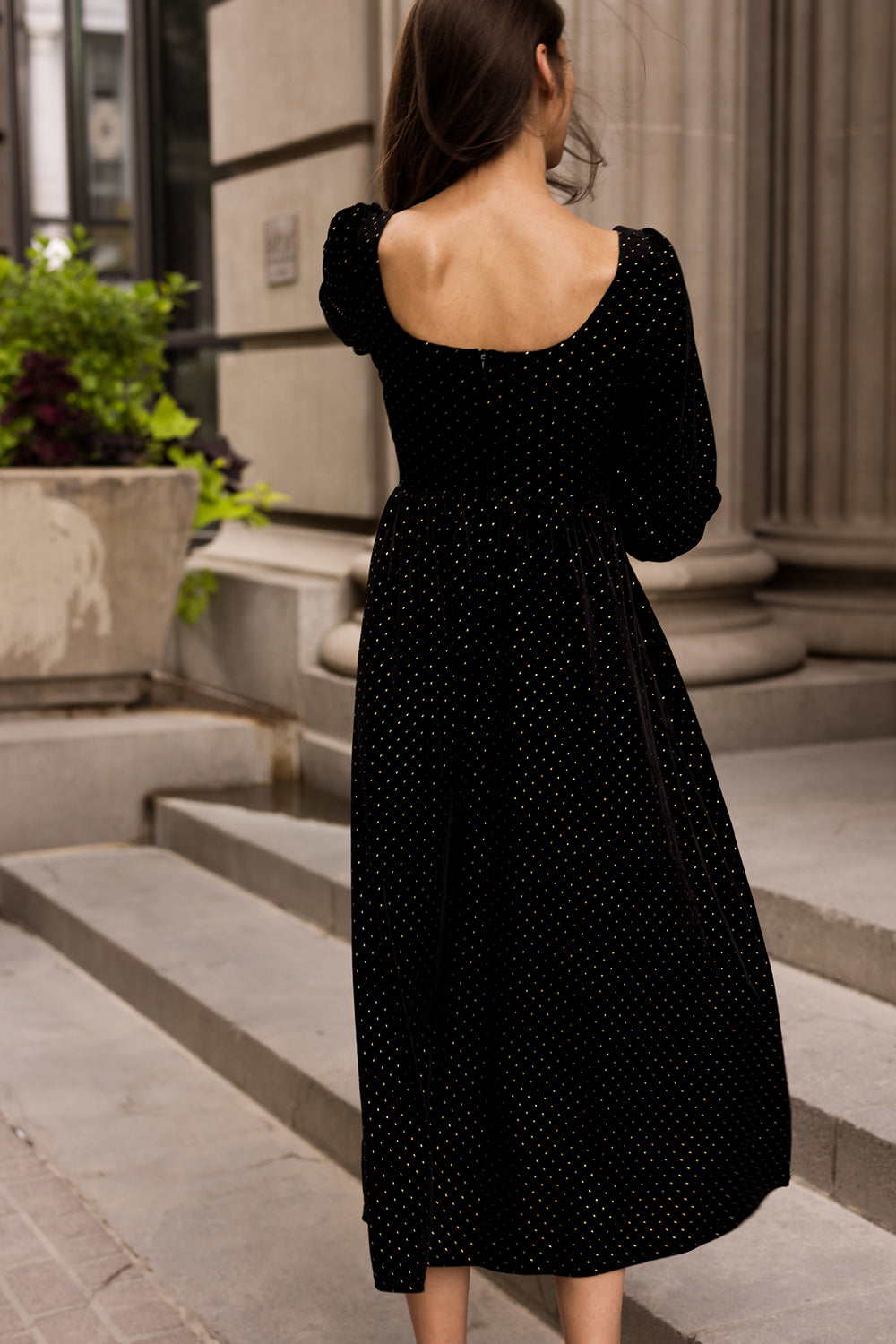 A woman with long brown hair, wearing the Ivy City Co Kingston Dress in Black featuring polka dots and long sleeves, stands on stone steps with her back to the camera in an urban setting near large columns and a planter.