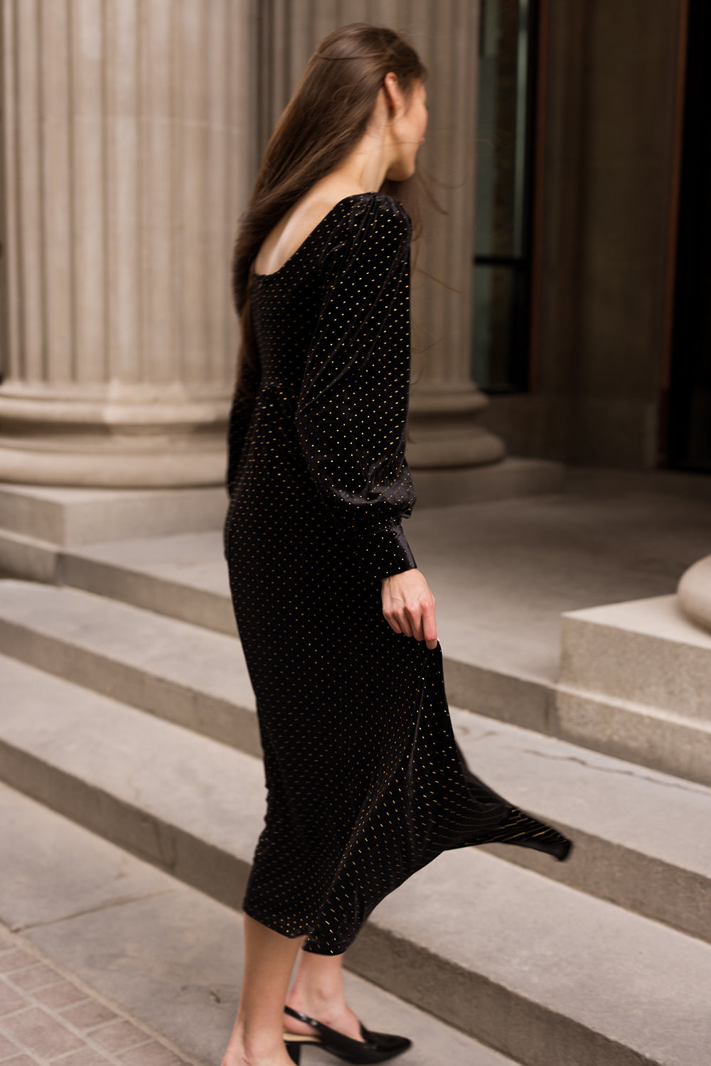A woman wearing the Ivy City Co Kingston Dress in Black—which features small white dots and long sleeves—walks down stone steps outside a building with large columns.