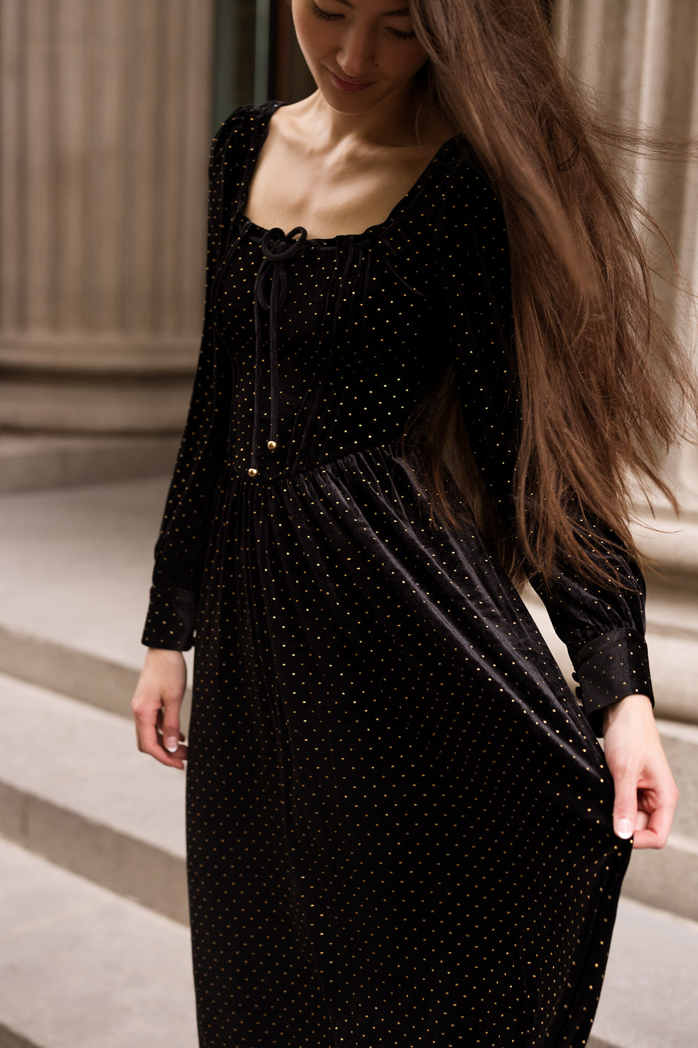 Wearing the Kingston Dress in Black by Ivy City Co—a long-sleeved dress adorned with gold polka dots—a woman stands gracefully on stone steps, gently holding her skirt with one hand and gazing down with a soft smile.