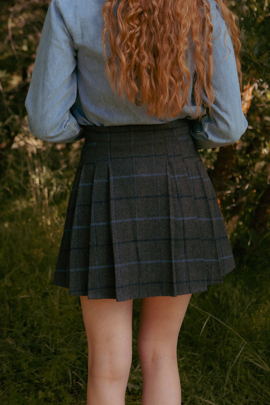A person with long, wavy auburn hair stands outdoors in front of greenery, wearing a light denim shirt and the Ivy City Co Kinley Skirt in Charcoal Plaid. Their back is to the camera.