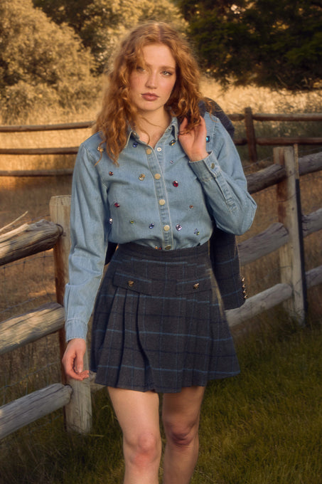 A woman with curly red hair wears the Ivy City Co Kinley Skirt in Charcoal Plaid and a denim shirt with embroidered details as she walks outdoors near a wooden fence, surrounded by trees and grass.