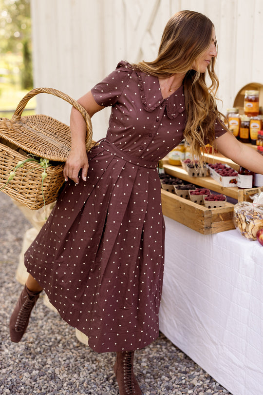 At an outdoor market, a woman in Ivy City Co’s Kit Dress in Warm Cocoa Dainty Dot—crafted from cotton with an easy, nursing-friendly design—holds a large basket and reaches for jars on a table filled with fruit and preserves.
