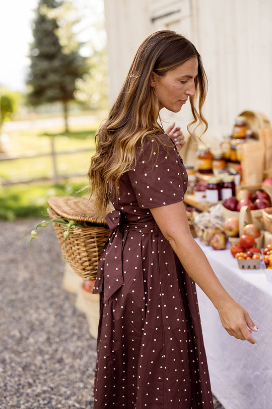 A woman wearing the Ivy City Co Kit Dress in Warm Cocoa Dainty Dot—a comfortable, nursing-friendly cotton midi—carries a wicker basket as she shops at an outdoor farmers market, browsing tables filled with fresh fruit and jars of jam.