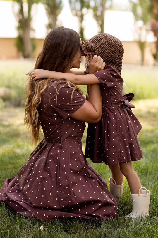 A mother and young daughter share a tender moment outdoors, both wearing the Ivy City Co Kit Dress in Warm Cocoa Dainty Dot. The girl completes her look with a straw hat and rain boots as they embrace face-to-face on a green lawn, showcasing how these nursing-friendly dresses blend style and comfort effortlessly.