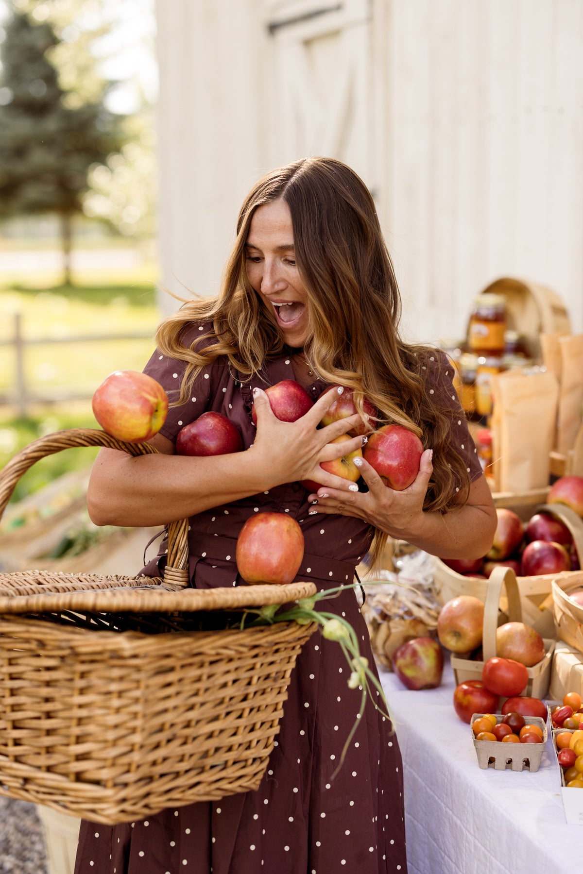A woman in the Ivy City Co Kit Dress in Warm Cocoa Dainty Dot smiles as she tries to hold several apples in her arms. She stands beside a table filled with fresh produce and baskets, outdoors near a white building. The nursing-friendly Kit Dress adds comfort and charm to this delightful scene.