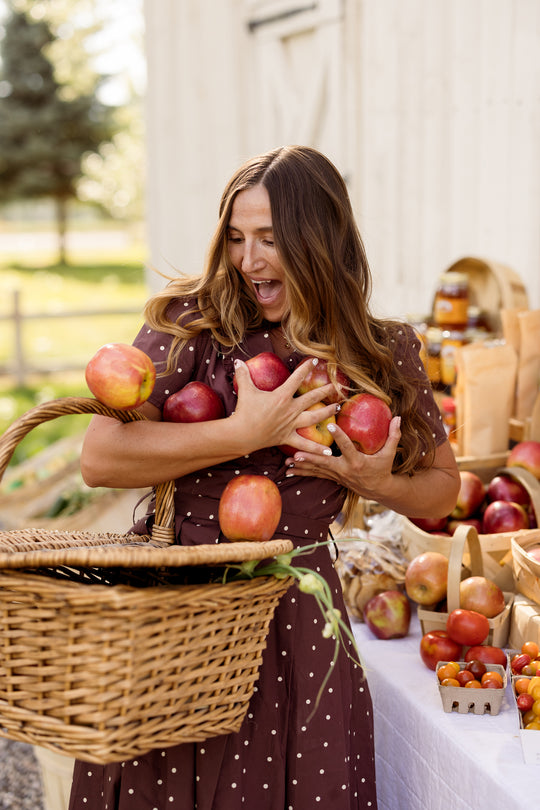A woman in the Ivy City Co Kit Dress in Warm Cocoa Dainty Dot smiles as she tries to hold several apples in her arms. She stands beside a table filled with fresh produce and baskets, outdoors near a white building. The nursing-friendly Kit Dress adds comfort and charm to this delightful scene.