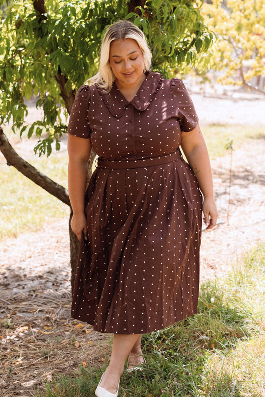 A blonde woman stands outside beneath a leafy tree, smiling and looking down as sunlight filters through the branches onto the grass. She wears Ivy City Co’s Kit Dress in Warm Cocoa Dainty Dot—a nursing-friendly, cotton midi dress with white polka dots—and white shoes.