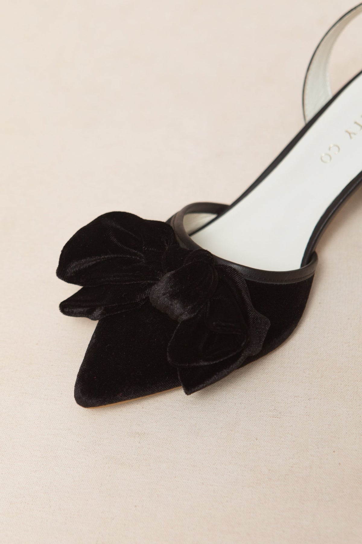 A close-up of the Eloise Bow Heel in Black by Ivy City Co showcases its elegant black velvet finish, a statement matching bow on the pointed toe, and a classic sling back design, all beautifully displayed against a neutral background.