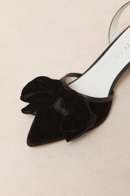 A close-up of the Eloise Bow Heel in Black by Ivy City Co showcases its elegant black velvet finish, a statement matching bow on the pointed toe, and a classic sling back design, all beautifully displayed against a neutral background.