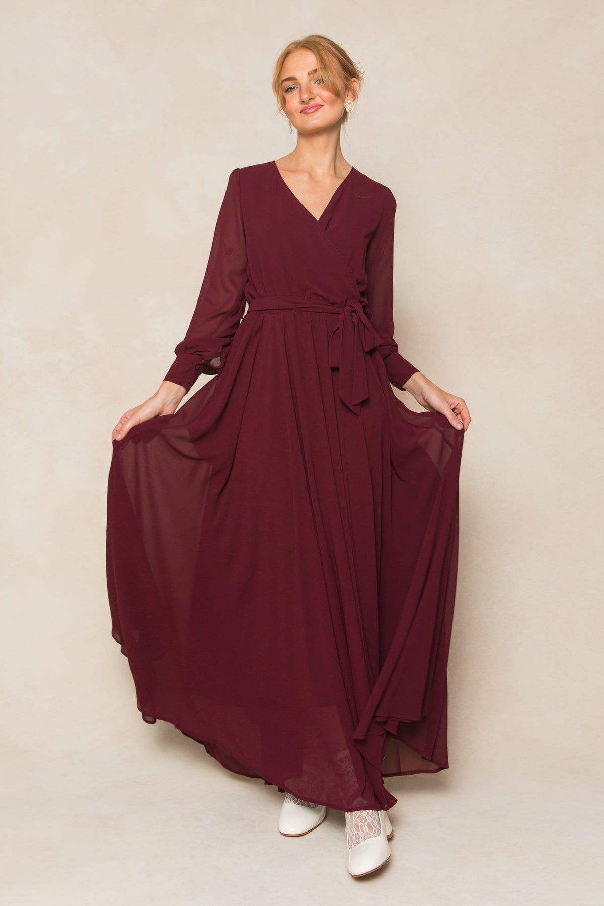A woman poses holding out the sides of the flowing skirt of the Andie Dress in Wine Chiffon by Ivy City Co. The long-sleeved maxi dress features a waist tie, and she styles her light hair up while wearing white boots.