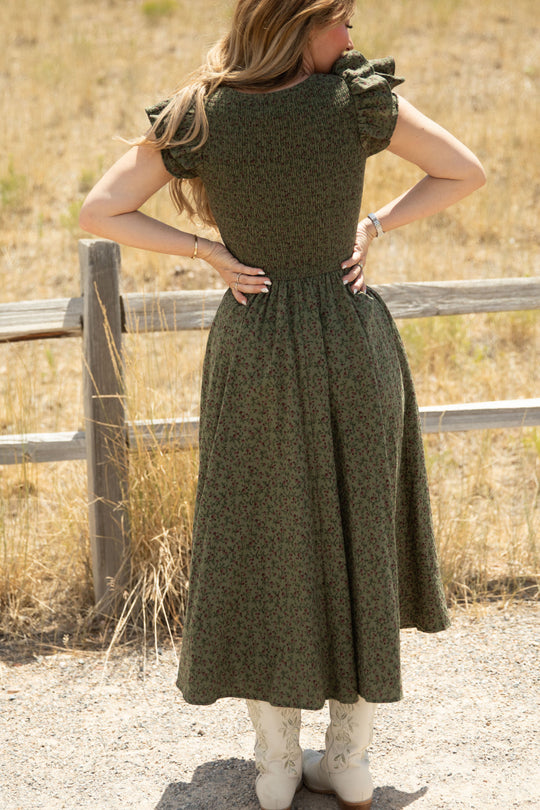 A woman with long hair stands outdoors, facing away. She wears the Ivy City Co Hattie Dress in Woodland Floral—a green floral style featuring a smocked bodice, ruffled sleeves, and white cowboy boots. A wooden fence and dry grass fill the background, emphasizing the dress’s maternity-friendly design.