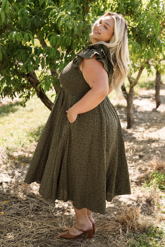 A woman in the Ivy City Co Hattie Dress in Woodland Floral, featuring a green ruffled design and smocked bodice, stands outdoors near trees, smiling with one hand in her pocket. Sunlight filters through the leaves, highlighting her blonde hair and the flowy, maternity-friendly dress.