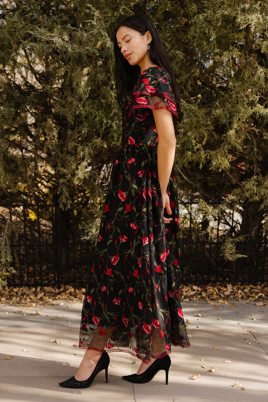 A woman stands sideways on a sidewalk surrounded by green trees and fallen leaves, wearing the Ivy City Co Flora Dress in Romantic Roses—a black dress adorned with red floral patterns and a black mesh overlay.