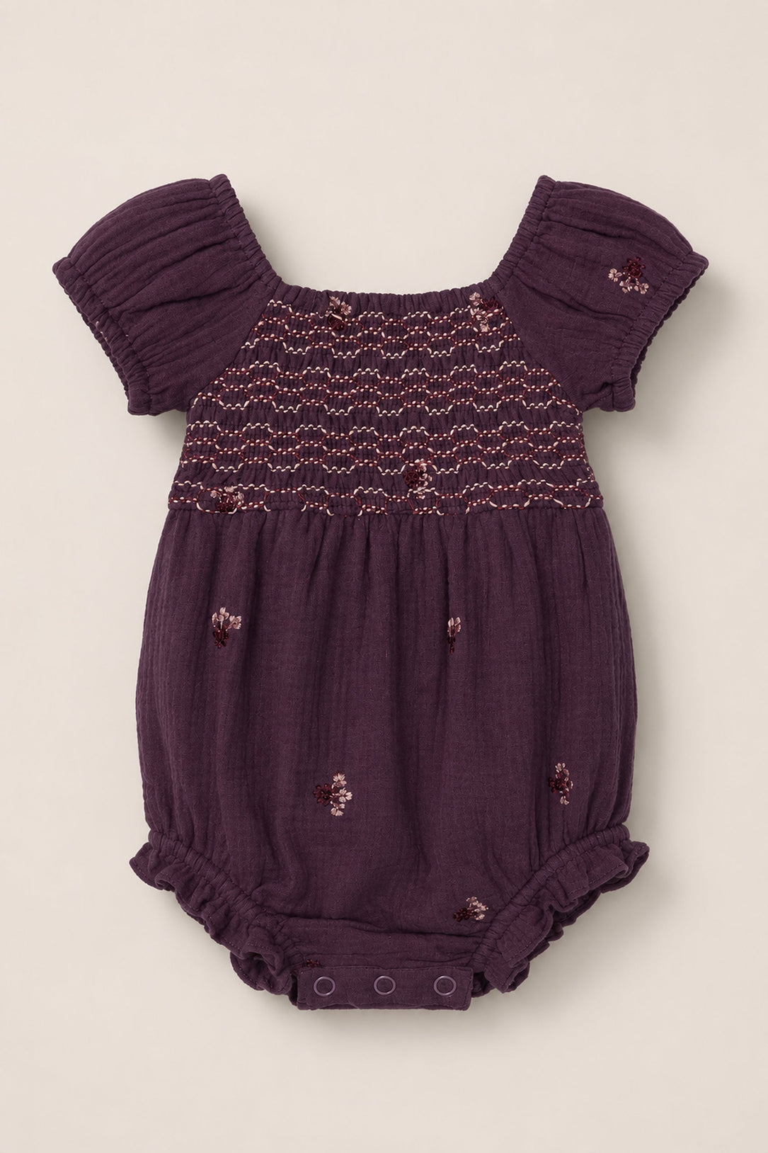 The Baby Acantha Romper in Aubergine Embroidered Gauze by Ivy City Co features short puff sleeves, a smocked chest, and delicate embroidered floral details. Designed for comfort, this purple cotton romper includes elastic leg openings and snap buttons at the bottom for easy diaper changes.