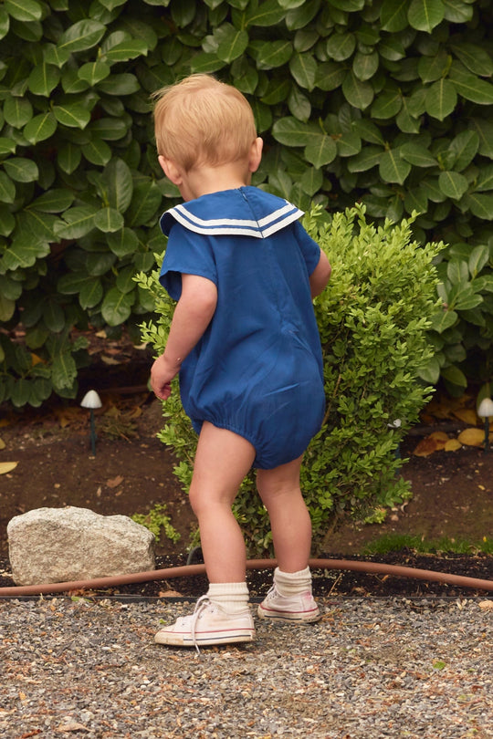 A toddler wearing the Ivy City Co Baby Sailor Romper in Navy and pink shoes stands outdoors on gravel, facing away from the camera near green bushes.
