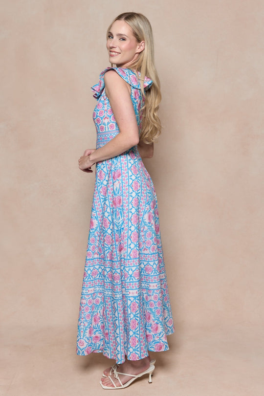 A woman with long blonde hair wearing the Samara Dress in Thistle Flower Mosaic Florise by Ivy City Co and white heels stands sideways and smiles in front of a beige background.