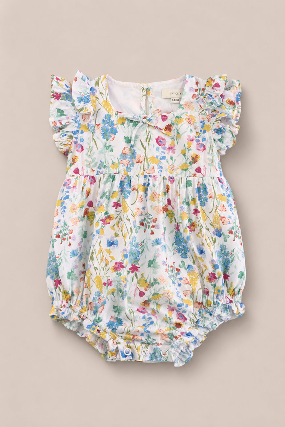 The Baby Ana Romper in Floral Fields by Ivy City Co features a colorful floral print, ruffled short flutter sleeves, and elastic leg openings, shown on a beige background—making it the perfect baby floral romper for summer.
