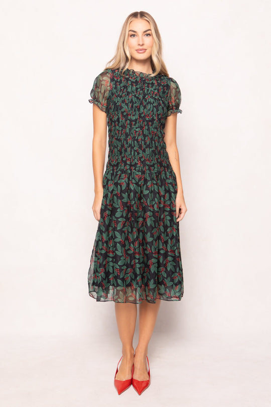 A woman wears a black, maternity-friendly Hayden Dress in Berries featuring a smocked bodice, short puff sleeves, and a green leaf and red berry print, paired with red pointed-toe heels.