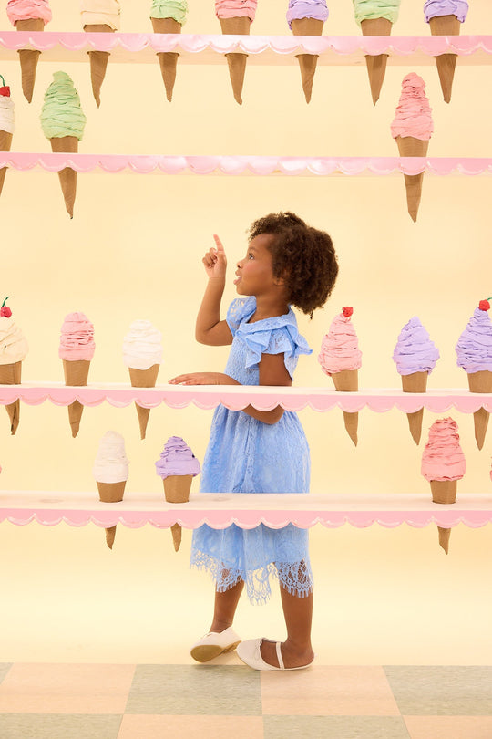 A young girl wearing the Ivy City Co Mini Hattie Dress in Cornflower Blue Lace points excitedly at colorful ice cream cones displayed on shelves against a pastel yellow background.