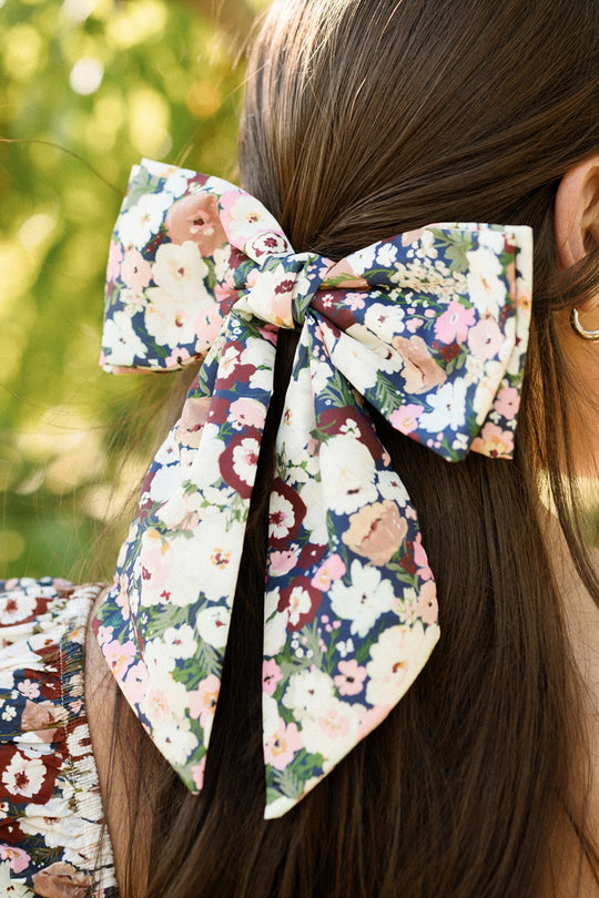 A close-up of a woman’s brown hair styled with the Ivy City Co Hair Bows in Midnight Garden, featuring a large, floral-patterned fabric bow. She wears a matching Ivy City Co floral top and gold hoop earrings against a sunlit green background.