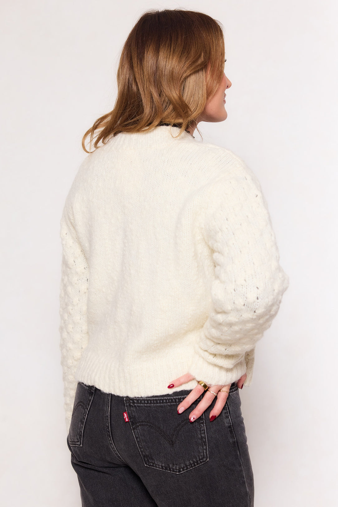 A person with light brown hair faces away, wearing the Ivy City Co Larissa Sweater in Cream and black jeans with a visible red tag on the back pocket against a plain white background.