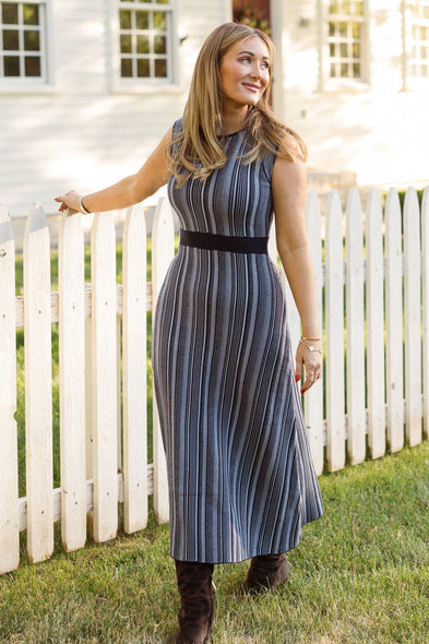 A woman, smiling and looking to the side, stands by a white picket fence on a sunny day wearing the sleeveless Laurel Dress in Midnight by Ivy City Co and brown boots. The striped blue dress is maternity friendly, offering both style and comfort for every stage.