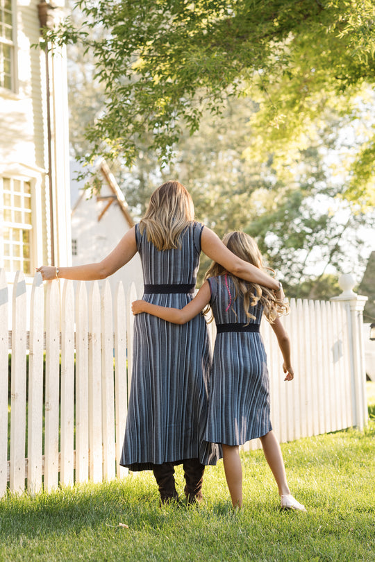 Two people, one adult and one child, both dressed in Ivy City Co’s Laurel Dress in Midnight—one of them styled with a cozy sweater knit—walk side by side through a grassy yard next to a white picket fence, their arms around each other as sunlight filters through the trees.