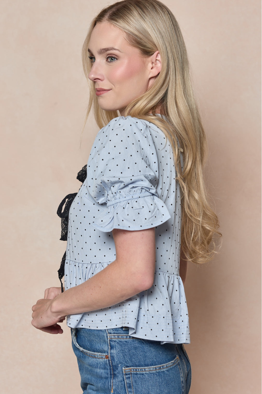 A woman with long blonde hair stands sideways against a light beige background, wearing the Ivy City Co Lavinia Top in Light Blue with short ruffled sleeves and polka dots, paired with blue jeans.