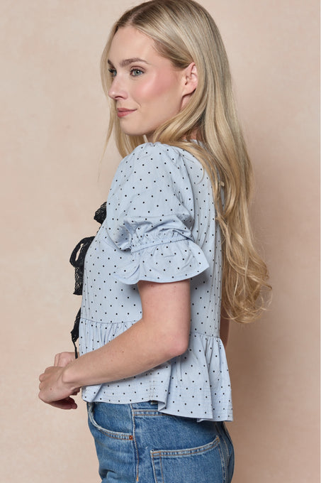 A woman with long blonde hair stands sideways against a light beige background, wearing the Ivy City Co Lavinia Top in Light Blue with short ruffled sleeves and polka dots, paired with blue jeans.