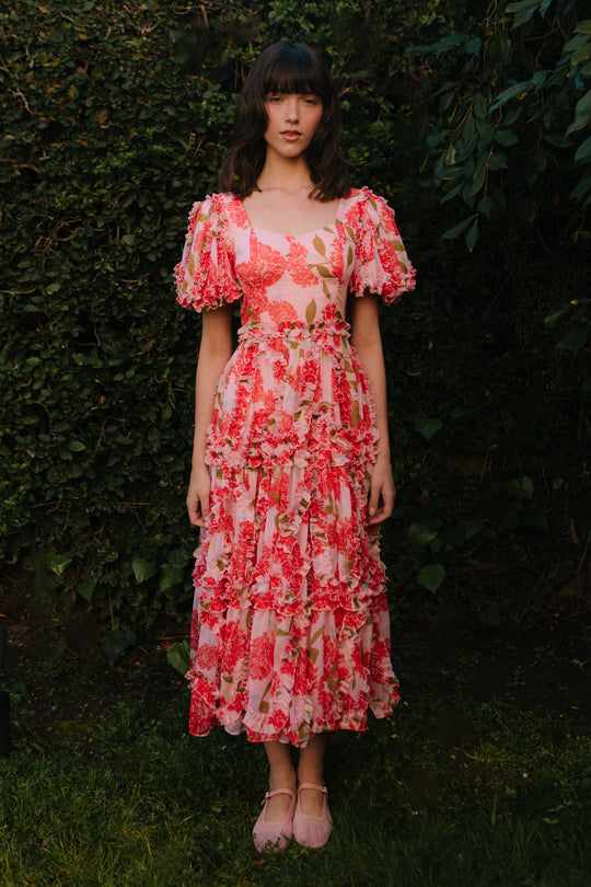 A woman stands outdoors against lush greenery, wearing the Ivy City Co Layla Dress in Pink Floral—a chiffon midi dress with ruffle details and puff sleeves—paired with light pink shoes, complementing her dark hair and neutral expression.