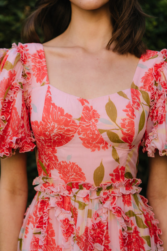 A woman outdoors wears the Ivy City Co Layla Dress in Pink Floral, featuring bold red patterns and delicate ruffled short sleeves, shown from shoulders to waist.