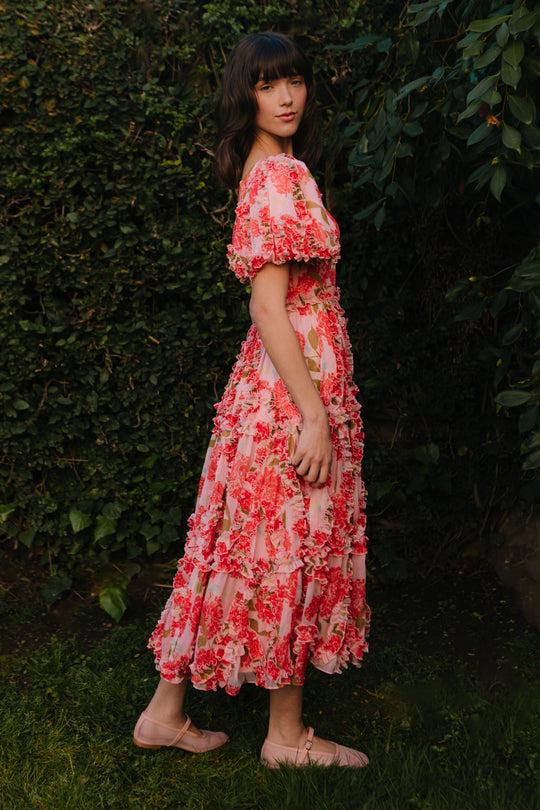 A woman stands sideways in a garden of lush green foliage, wearing Ivy City Cos Layla Dress in Pink Floral with ruffle detailing and light pink shoes.