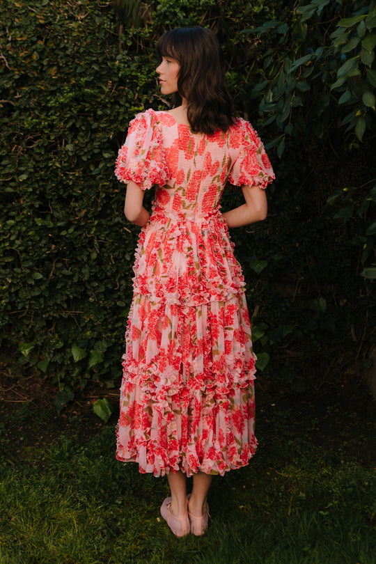 A woman with shoulder-length brown hair stands on grass facing away, wearing the Ivy City Co Layla Dress in Pink Floral, featuring puffed sleeves, ruffle detailing, and a fitted waist, in front of lush green foliage.