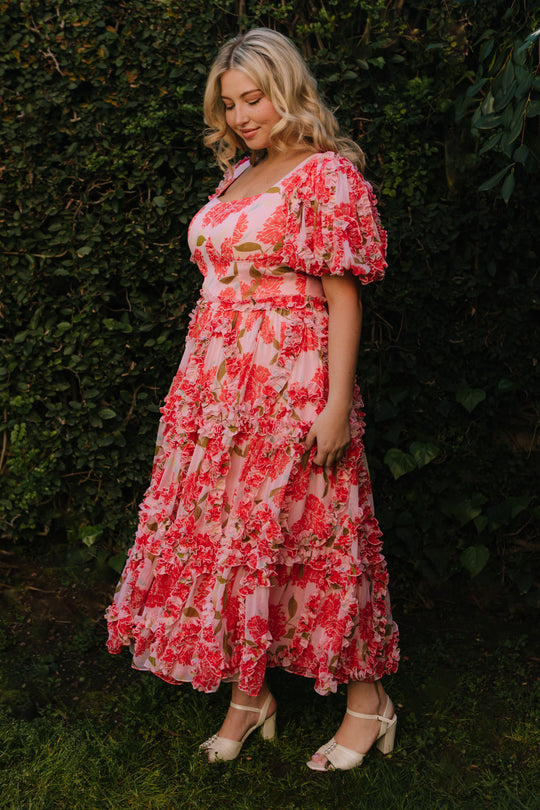 A woman with blonde hair stands on grass amid greenery, wearing the Ivy City Co Layla Dress in Pink Floral—a chiffon midi dress with ruffle detailing—paired with white heeled sandals. She faces slightly to the side with a calm expression.