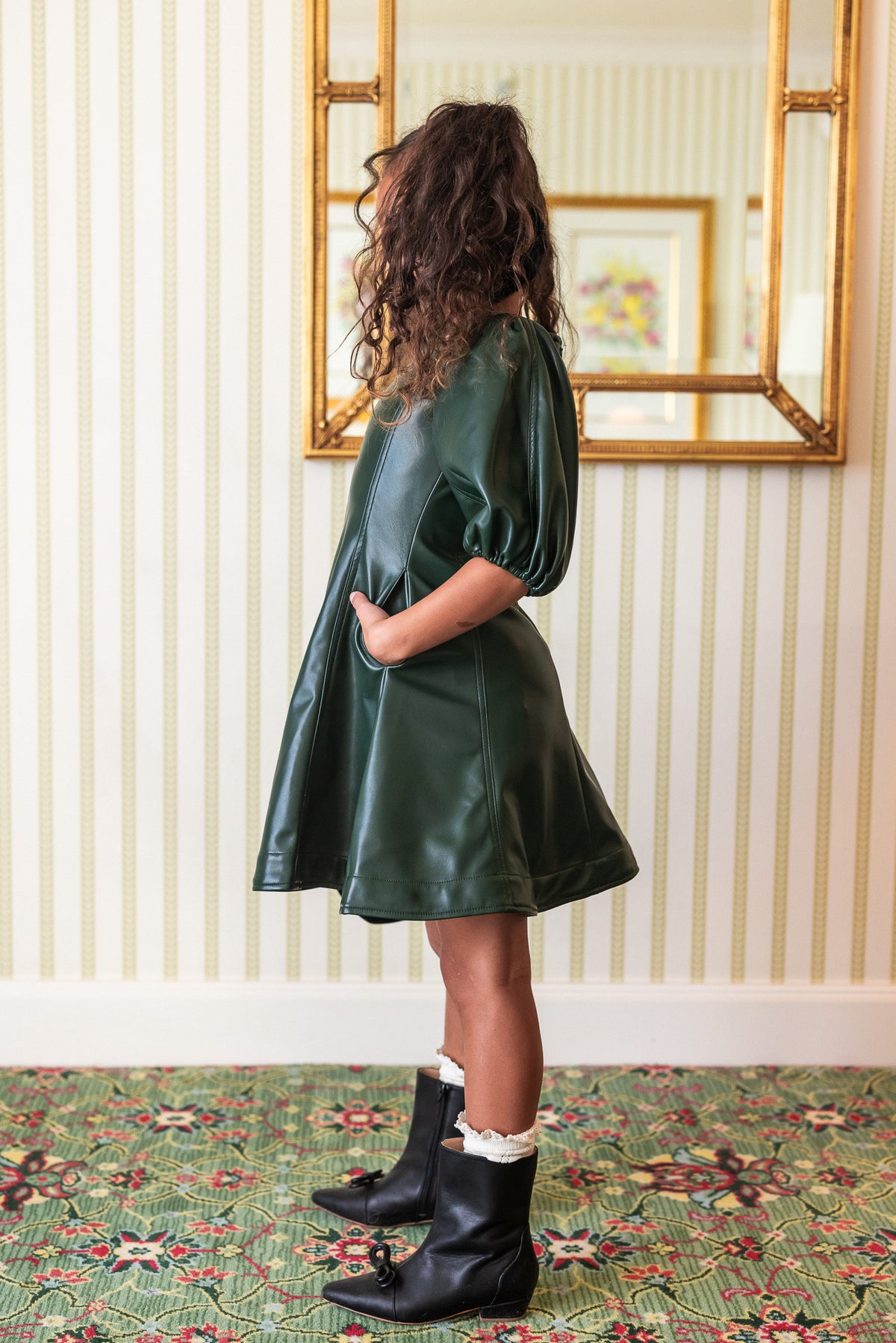 A girl wearing a green vegan leather mini dress with a v neckline and long balloon sleeves. She is also wearing white tights and black boots, carrying a small black purse.
