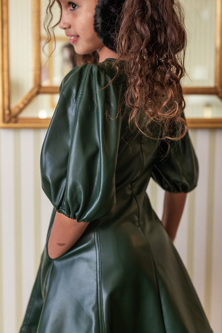 A girl wearing a green vegan leather mini dress with a v neckline and long balloon sleeves. She is also wearing white tights and black boots, carrying a small black purse.