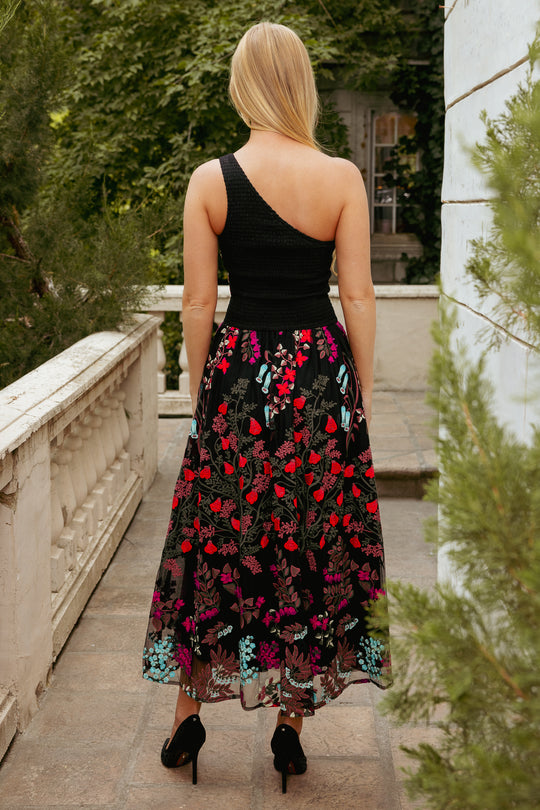 woman in multicolored one shoulder sleeve dress with embroidery standing on a patio with greenery behind